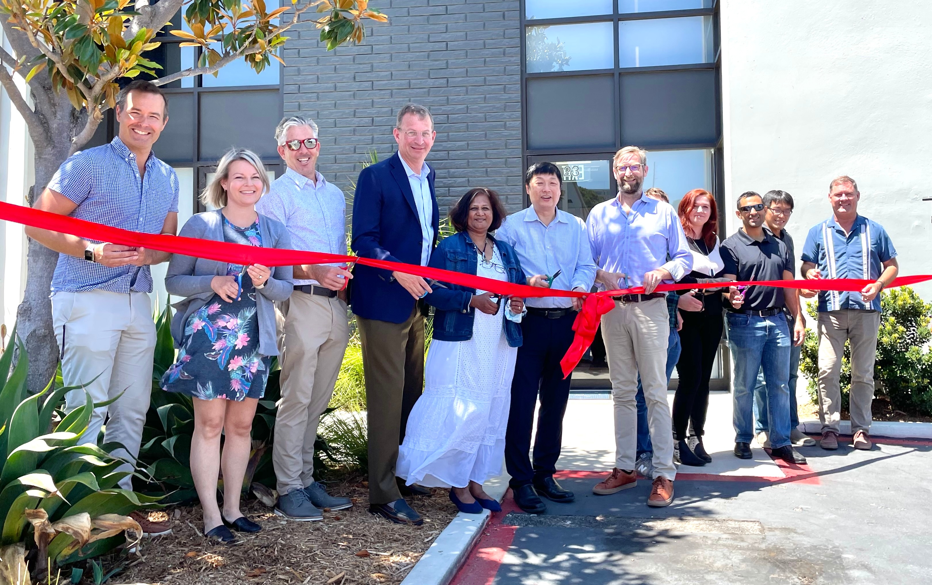  Group of people outdoors on a sunny day in San Diego holding a red ribbon in front of a building, as at a ribbon-cutting event