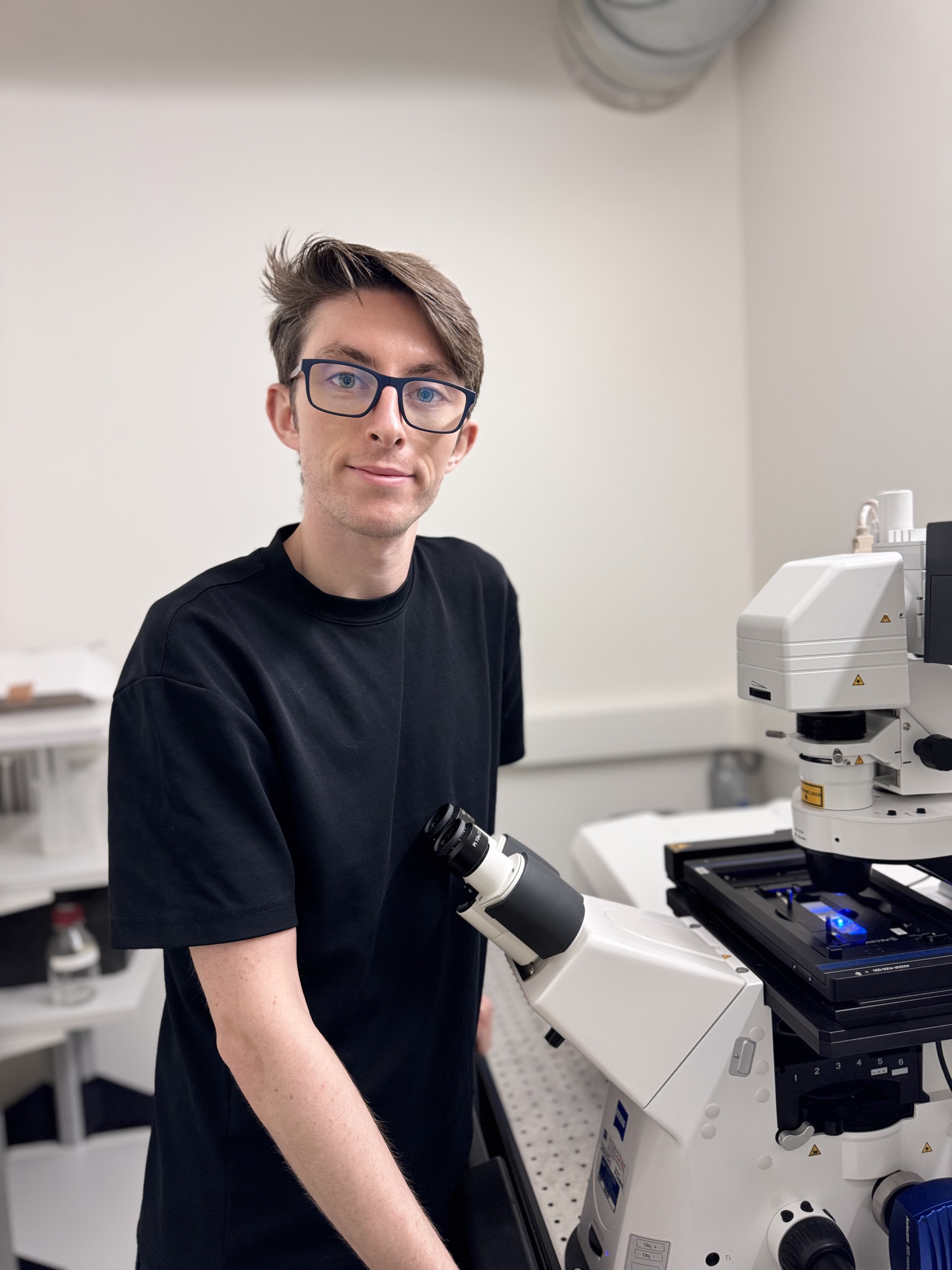 A person wearing glasses and a black shirt is standing by a Zeiss microscope in a laboratory setting.